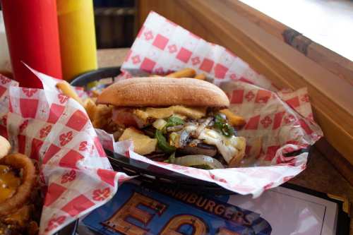 A close-up of a burger with toppings in a basket, surrounded by condiments and fries on a table.
