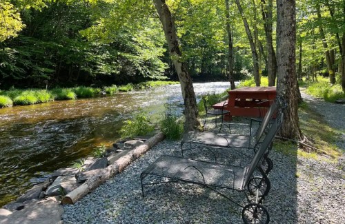 A peaceful riverside scene with two chairs and a picnic table surrounded by trees and a gently flowing stream.