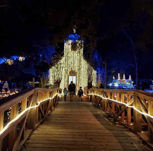 A family walks across a wooden bridge towards a beautifully lit church surrounded by festive decorations.