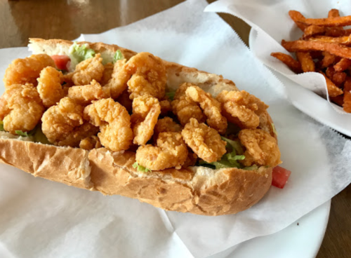 A shrimp po' boy sandwich filled with crispy shrimp, lettuce, and tomato, served with a side of sweet potato fries.