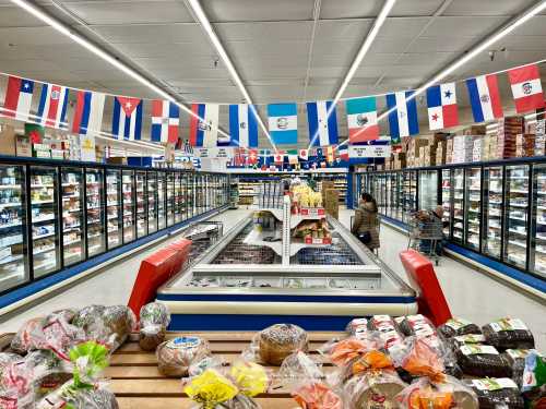 A grocery store aisle with freezers, international flags hanging above, and various baked goods in the foreground.