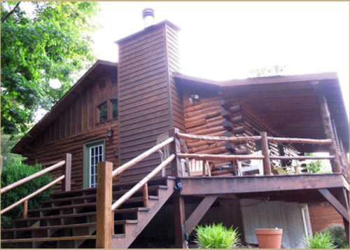A rustic log cabin with a chimney, wooden deck, and steps, surrounded by greenery.