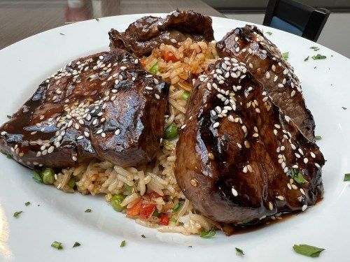 A plate of glazed beef slices on a bed of fried rice, garnished with sesame seeds and mixed vegetables.