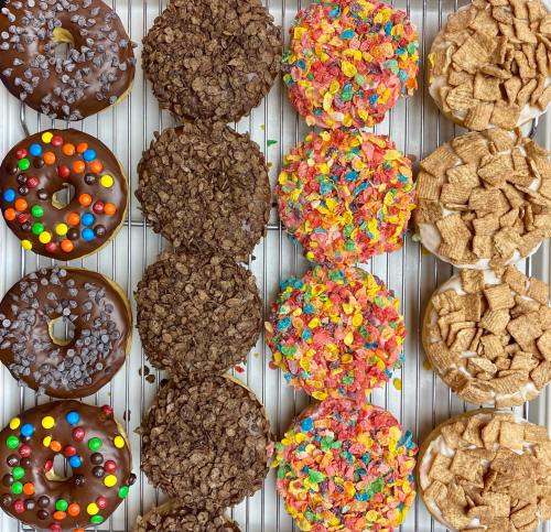 A variety of colorful donuts topped with chocolate, cereal, and candy, arranged neatly on a metal tray.