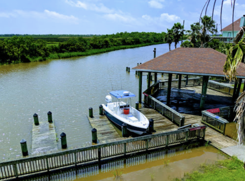 A boat docked at a wooden pier beside a calm river, surrounded by lush greenery and a clear blue sky.
