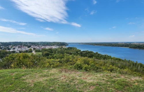 A scenic view of a river winding through green trees, with a small town visible in the distance under a blue sky.