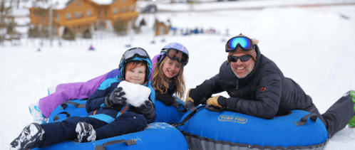 A man and two children smile while sitting on blue snow tubes in a snowy landscape, ready for winter fun.