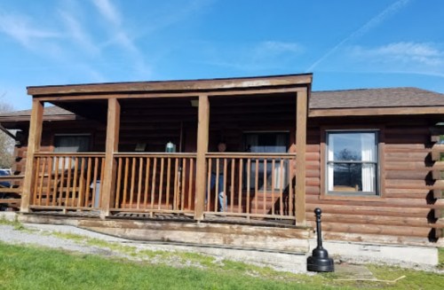 A rustic log cabin with a porch, surrounded by grass and blue sky, featuring large windows and a wooden railing.