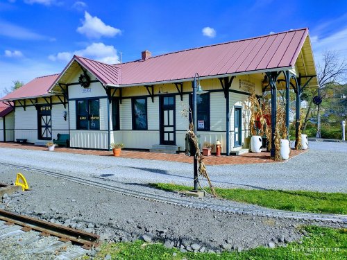 A charming train station with a red roof, surrounded by gravel and decorative plants, under a blue sky.