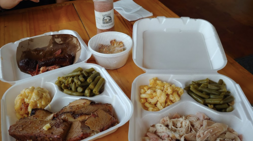 Two takeout containers with barbecue brisket, chicken, green beans, macaroni, and sides on a wooden table.