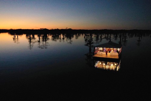 A serene sunset over a calm lake, featuring a lit pavilion surrounded by silhouetted trees.