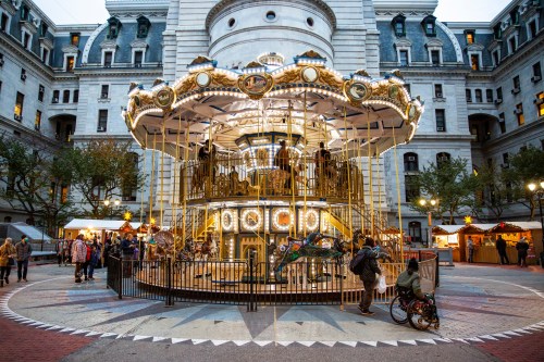 A beautifully lit carousel in a festive square, surrounded by people and holiday decorations.