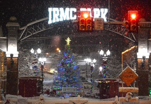 A snowy scene at IRMC Park featuring a decorated Christmas tree and festive lights under a glowing archway.