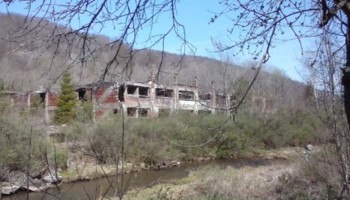 Abandoned building surrounded by trees and a river, set against a backdrop of rolling hills under a clear blue sky.