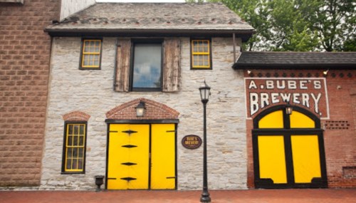 Historic brewery building with yellow doors and windows, featuring stone and brick walls, surrounded by greenery.