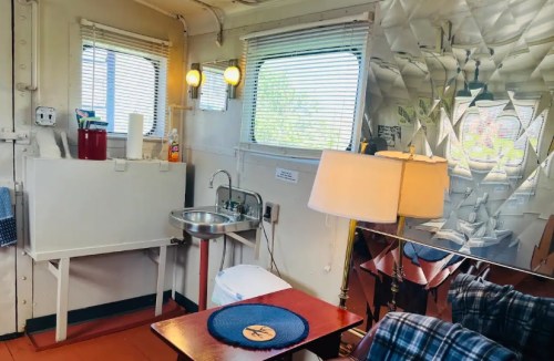 Interior of a small room featuring a sink, lamp, table, and reflective wall, with natural light from windows.