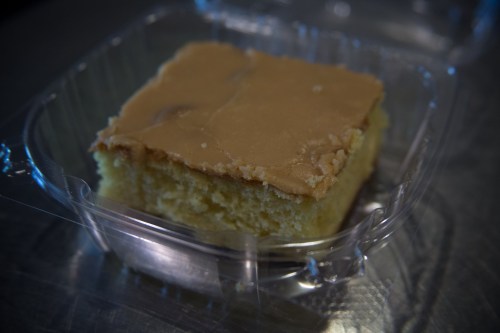 A square piece of cake with a smooth, light brown frosting in a clear plastic container.