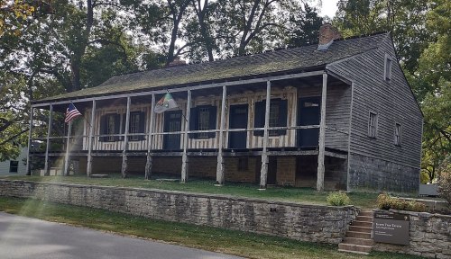 Historic wooden building with a porch, flags, and stone foundation, surrounded by trees and grass.