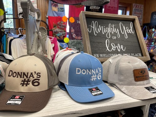 Three caps with "Donna's #6" in different colors, displayed on a table next to a sign that reads "Almighty God of love."