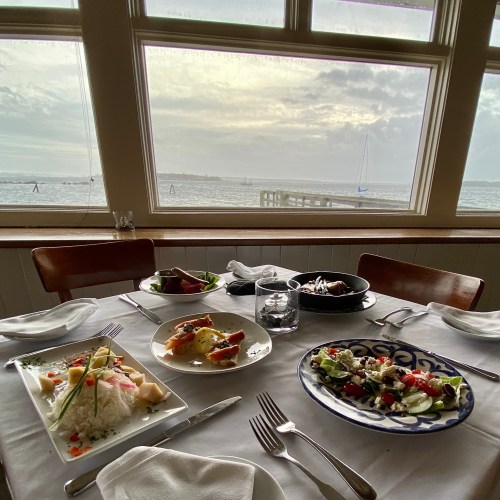 A dining table set with various dishes, overlooking a scenic view of water and boats through large windows.