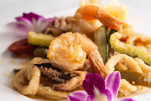 A close-up of a seafood dish featuring shrimp, calamari, and vegetables, garnished with purple orchids.