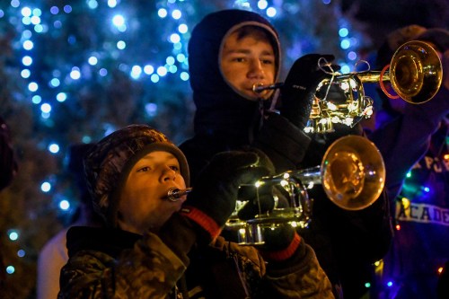 Two boys play trumpets, surrounded by festive blue lights, wearing winter clothing and gloves.