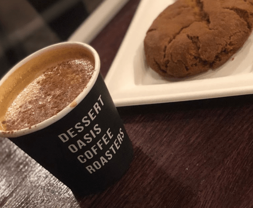 A coffee cup from Dessert Oasis Coffee Roasters next to a large cookie on a white plate.