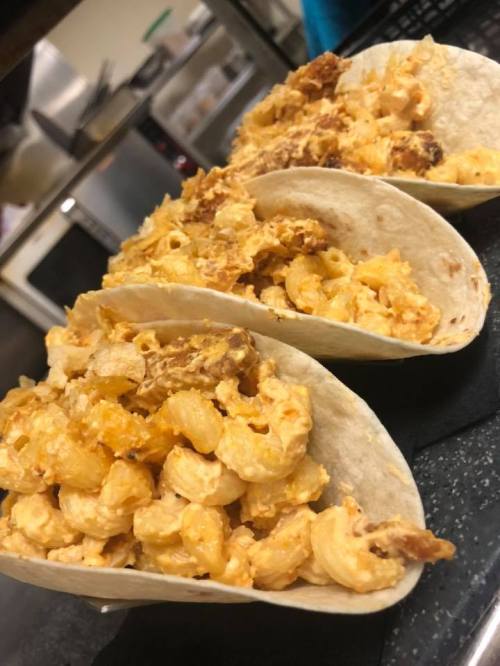 Three tacos filled with creamy macaroni and cheese, topped with crispy bits, served on a black surface.