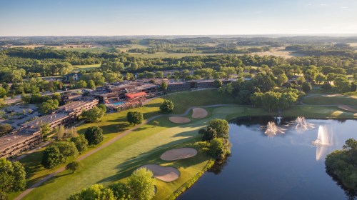 Aerial view of a golf course with a pond, fountain, and lush greenery, alongside a resort and rolling hills in the background.