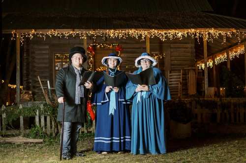 A group of three carolers in festive attire stand outside a decorated house with holiday lights.
