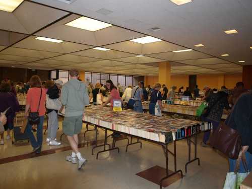 A crowded indoor book fair with people browsing tables filled with books. Brightly lit space with yellow walls.