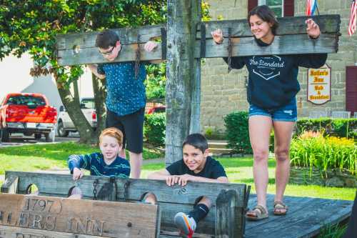 Four children play in a wooden stocks display outside a historic inn, smiling and enjoying the sunny day.