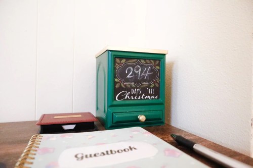 A green countdown calendar shows "294 DAYS 'TIL CHRISTMAS" next to a guestbook and a pen on a wooden table.