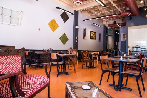 A cozy café interior with various wooden tables and chairs, colorful wall art, and vintage furniture.