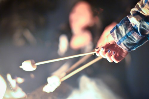 A hand holding skewers with marshmallows over a campfire, with a blurred figure in the background.