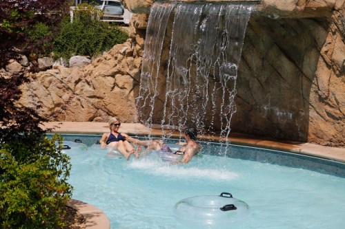 A woman and a man relax in a pool under a waterfall, surrounded by rocks and greenery.