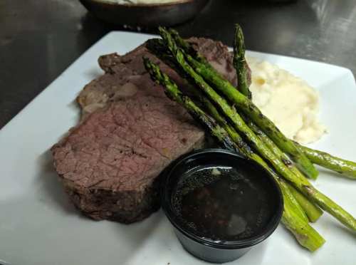 A plate with sliced roast beef, asparagus, mashed potatoes, and a small cup of sauce.