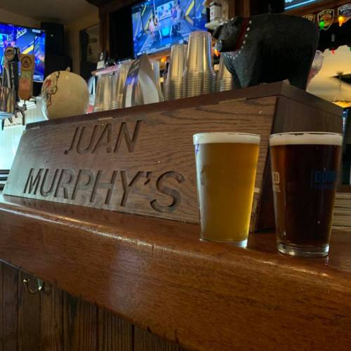 Two glasses of beer sit on a wooden bar with the name "JUAN MURPHY'S" carved into it, surrounded by bar decor.