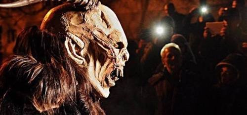 A person in a grotesque mask with horns, surrounded by an audience holding phones, in a dimly lit setting.