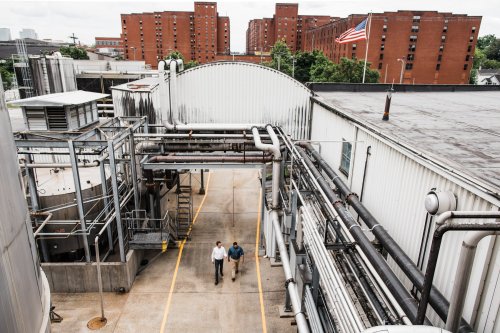 Two men walk along a concrete path between industrial buildings and pipes, with a flag and red brick buildings in the background.