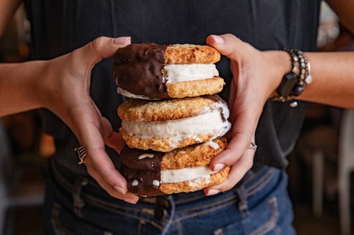 A person holds three stacked ice cream sandwiches, two with chocolate coating and one with vanilla.