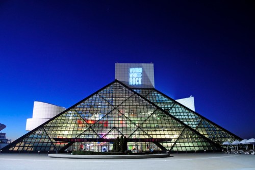 The Rock and Roll Hall of Fame at night, featuring a glass pyramid structure and a projection reading "Women Who Rock."