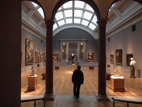 A person walks through an art gallery, surrounded by paintings and sculptures under a skylit ceiling.