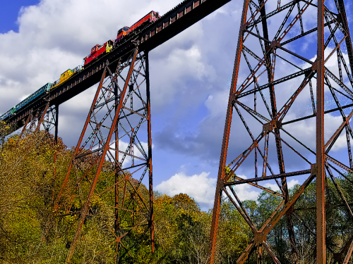 A colorful train crosses a tall, rusted bridge surrounded by trees under a partly cloudy sky.