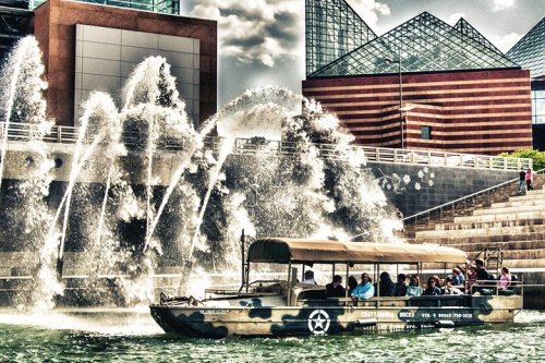 A boat with passengers navigates a waterway, surrounded by dramatic fountains and modern buildings.
