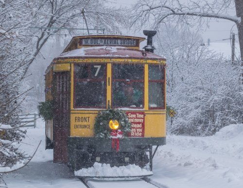 A vintage yellow tram decorated with wreaths travels through a snowy landscape, surrounded by trees and falling snow.