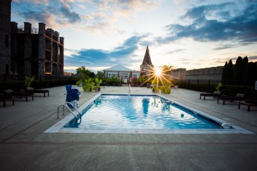 A serene pool area at sunset, surrounded by lounge chairs and greenery, with a historic building in the background.