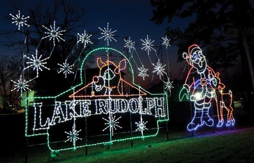 Colorful holiday lights display featuring "Lake Rudolph," a reindeer, Santa, and festive snowflakes against a twilight sky.