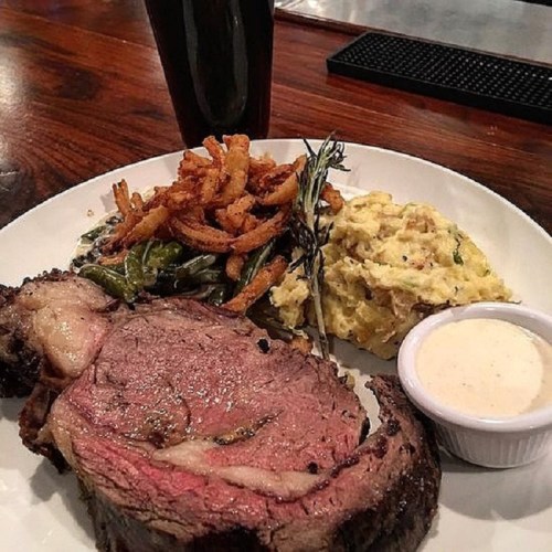 A plate featuring a slice of prime rib, creamy mashed potatoes, crispy onions, and sautéed greens, with a dipping sauce.
