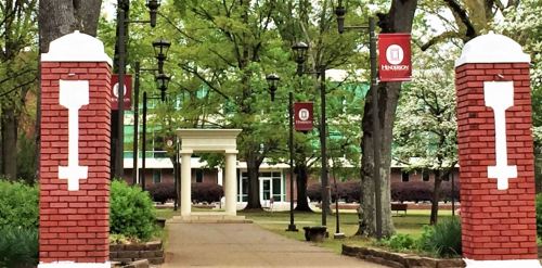 Entrance to a campus with brick pillars, banners, and trees lining the pathway leading to a building in the background.